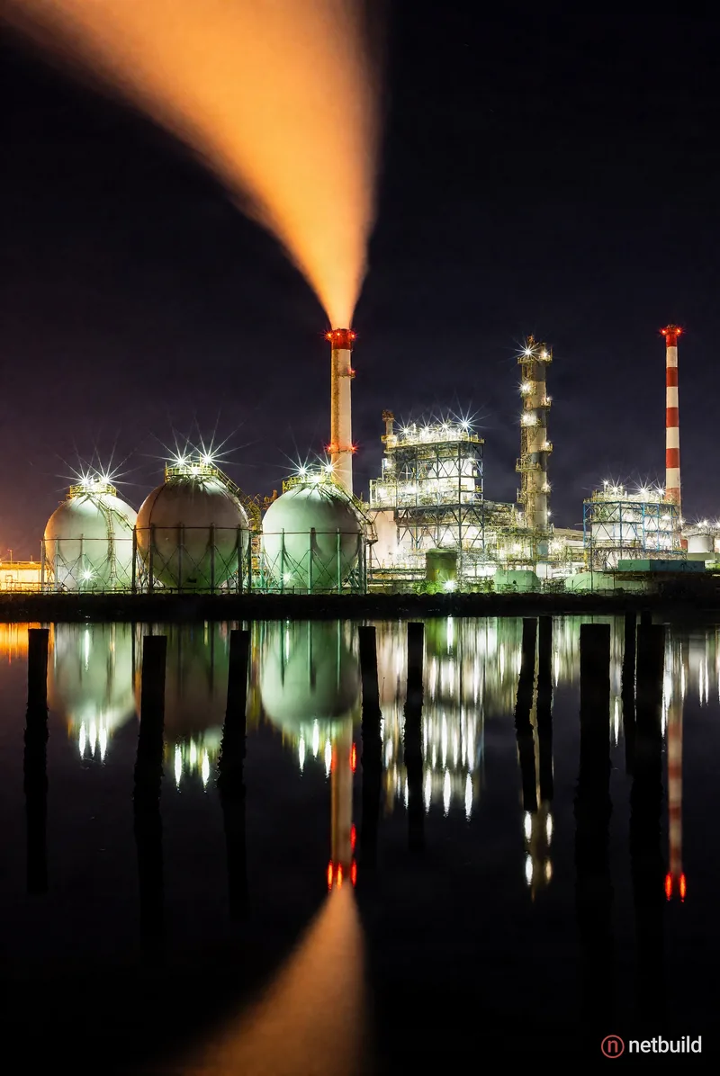 Crisp long exposure night photography of a glowing industrial facility across a glassy waterfront. Spherical storage tanks and towering smokestacks gl...